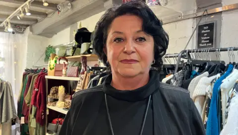 A woman with dark hair and a dark top is looking at the camera. Clothes are on coat hangers behind her left shoulder on the photo.