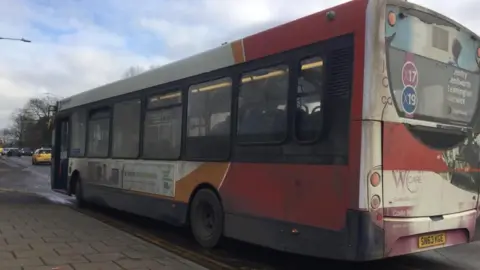 Single-decker bus with red, orange and white livery of Stagecoach.  The vehicle is quite dirty and is parked alongside a wide pavement.