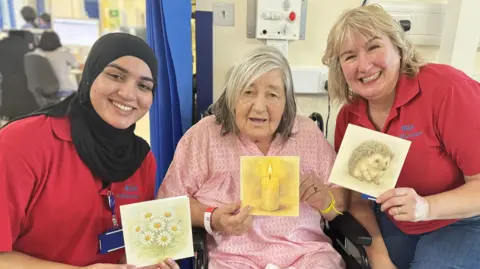 A woman in a pink gown sits in a chair holding a card of a burning candle. Either side of her are two women in red polos holding a card. All are smiling at the camera.