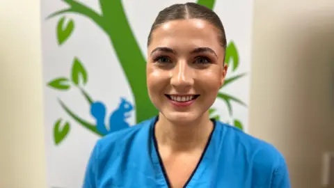 Physiotherapist Isla Lundie, a young medical professional, smiles, wearing her blue NHS scrubs. Her hair is tied back in a slick bun.