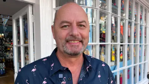 Phil Harrison/BBC Nick Wackett who works in the Pantiles. Nick is bald and is wearing a navy shirt with flamingos on it. He is stood outside a children's toy shop.