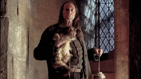 Murray Close/Getty Images An actor with long brown hair holds a cat in one hand and a lantern in the other inside a medieval castle, with stone walls and a leaded window behind him. He is dressed in a brown waistcoat, jacket and trousers and has a frown on his face.