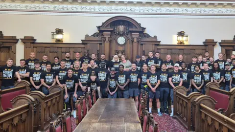 Port Vale at a civic reception with the Lord Mayor. The whole team is lined up wearing matching t-shirts. The Lord Mayor is at the back. They are in a grand civic building.