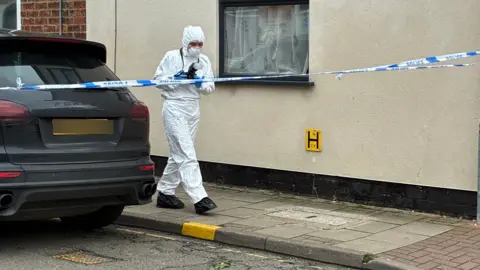 Jake Zuckerman/BBC A forensic police officer wearing a white protective suit stands on a pavement next to a dark vehicle. They are holding a camera and are working within a police cordon outside a building with a single window