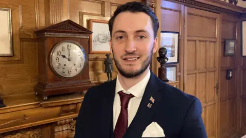 The image shows the interior of a traditional, wood‑panelled room with richly detailed décor. The space appears formal and historic, featuring carved wooden walls, framed pictures, and classic furnishings. A man is standing in the foreground wearing formal attire. He is dressed in a dark suit jacket with a white pocket square, a white shirt, and a dark red tie. The lapel of the jacket displays two small enamel pins—one featuring a small flag and another that appears to be a crest or emblem.