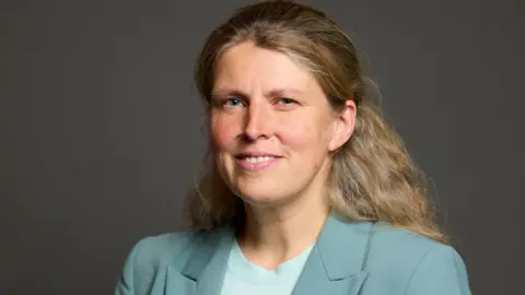 UK Parliament Official parliamentary photo portrait of Rachael Maskell. She has long wavy blonde hair and is wearing a pale blue suit and round-neck top.