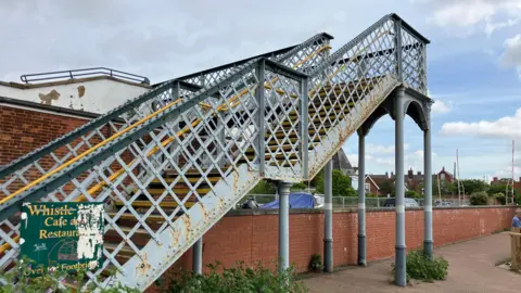 Steps leading up to a footbridge. The bridge has grey metal railings and yellow handrails, and some of its paint is discoloured.