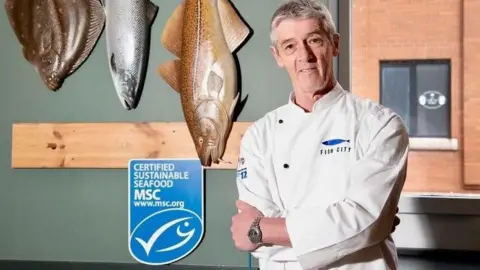 John Lavery John Lavery stands in a white uniform with a blue logo that says fish city. He is smiling. Behind him there are three large fish on the wall and a blue certified sustainable seafood sign.