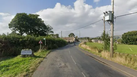 Google The entrance to the village of Romaldkirk. A sign of the village's name is on the  left of the road in the grass. Fields and trees can be seen on either side of the road. Stone-built houses can be seen in the village at a distance.