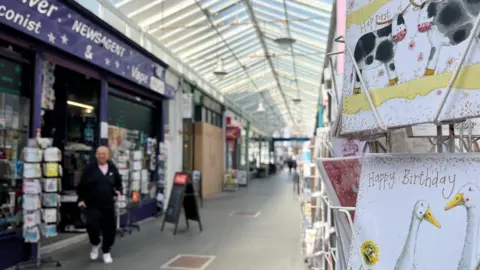 George Carden/BBC Inside the arcade which is lined with shops and chairs outside cafes. Some of the shops have post card holders outside and people are walking through the arcade.