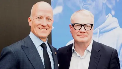 Two men stood smiling at the camera. Man on the left is bald and wearing a dark blue suit. Man on the left is wearing black framed glasses, a white shirt and black blazer.