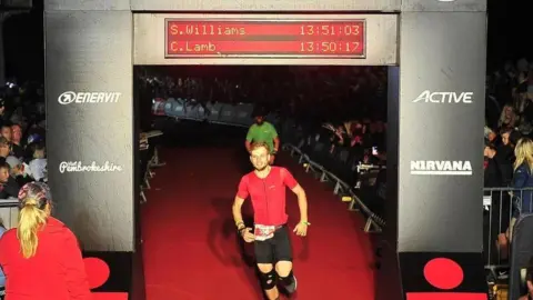 Liam Coffill Liam runs through a large black archway in an arena, which says Iron Man Wales. This photo is before his accident and he is wearing a red tshirt and black shorts. There are crowds sitting on either side.