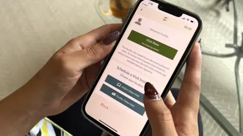 BBC News An image of a pair of hands holding a smartphone. The screen shows the homepage for an inmate visitation application, with buttons that say 'Visit Now', 'Online Video Visit' and 'Faculty Video Visit'. 