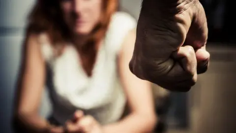 A man's fist coming from the top of the image into shot. In the background is a woman sitting down, who is blurred. 