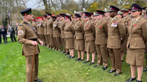 A regiment stand to attention in front of their commander during the remembrance service in Tidworth, Wiltshire. They are all wearing light brown uniforms with poppies attached and black and red caps. 