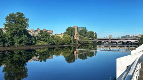 A bridge stretches over the still blue River Clyde in Glasgow. The sky is completely clear blue and buildings can be seen behind trees on the other side of the river from the viewer.