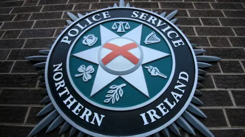 A close up of the PSNI emblem on a brick wall. It consists of a star shape in the centre, with the scales of justice, a harp, a torch, a leaf, a shamrock, and crown.