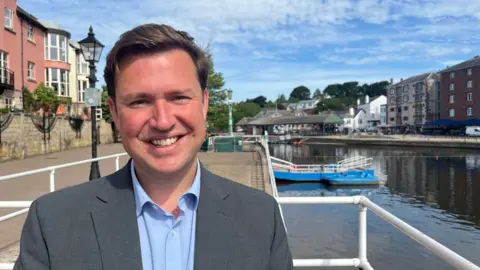 A smiling Steve Race in a blue shirt and a grey suit jacket standing on the quay in Exeter with the river to the right.
