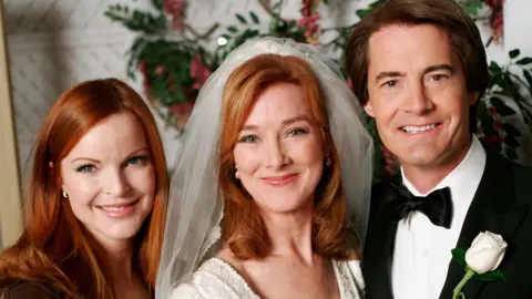 Getty Images Valerie Mahaffey is wearing a wedding dress and veil. she is standing next to a man in a tuxedo with a white rose boutonniere and a woman with red hair in a black dress.