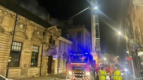 Michael Nagasaka/BBC A stone building ablaze at night, with black smoke coming from the roof. A fire engine sits in front of the building. A large fire truck with an extended ladder sprays water. The street is cordoned off with emergency crews present.