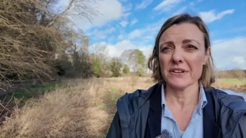A woman in a blue coat stands under blue skies next to the River Meon 