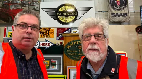 A recent seflie of David with John. David, on the left, wears a blue shirt and a red high vis vest. John, on right, wears a blue shirt, black jacket and a red high vis jacket. Both wear black metal glasses.