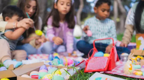 Getty Images Several children and one adult sit outside painting eggs. Some eggs sit on a box with others on a blanket.