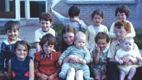GaelStair Project Twelve children smiling for the camera in 1970s clothing. Behind them is a paved footpath and a patch of grass alongside a brick house.
