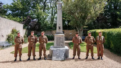 Lucie Stribrska The same six soldiers are seen stood either side of a war memorial in a garden with brown gravel and a large tree on a grassy area behind them.