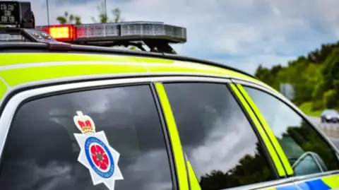 Lancashire Police A police car is parked facing a road. It has a sticker on the back window which reads Lancashire Constabulary. A blue sky and some trees can be seen in the distance over a road.