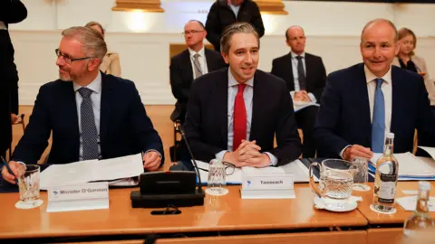 PA Media Three men - Roderic O'Gorman, Simon Harris and Micheál Martin - sit together at a large table. On the left, O'Gorman is wearing a suit and tie. He has grey hair, is wearing rectangle shaped glasses and is looking to the side. Harris sits in the middle and is mid-speech, wearing a suit and tie. Martin is sat on the right and smiles while wearing a suit and tie.