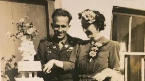 Hans Obma An old black and white photograph from the 1930s shows and man and woman cutting a white tiered cake on their wedding day. 