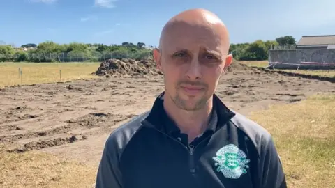 BBC Guernsey Football Association chief executive Gary Roberts, he has a bald head and stubble. He is wearing a blue top which included the white and green shield of the Guernsey FA.