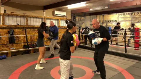 Two female boxers are in a ring sparring with male trainers.