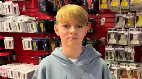 Keelan, 14, with blond hair wearing a grey hoodie standing in front of darts flights on sale in a shop.