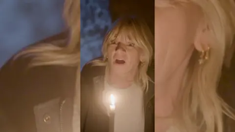 Zoe Ball has long blonde hair she is wearing a white top and a dark coat by candle-light. She looks shocked.