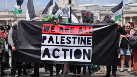 A banner for Palestine Action being held up reading 'We Are All Palestine Action' - it is being held up at what appears to be a protest, with a number of Palestine flags visible in the background 