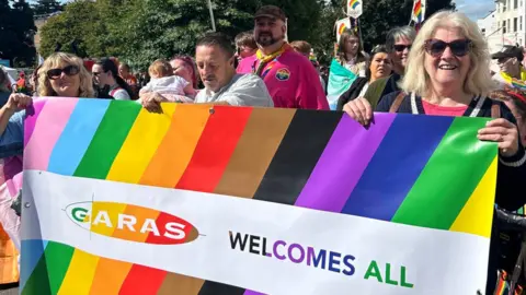 Adele Owen in a crowd of GARAS clients and supporters on a sunny day, holding up a rainbow-coloured banner saying GARAS welcomes all