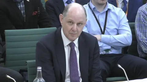 Sir Philip Barton sits in the committee room. he is wearing a suit and a purple patterned tie. 