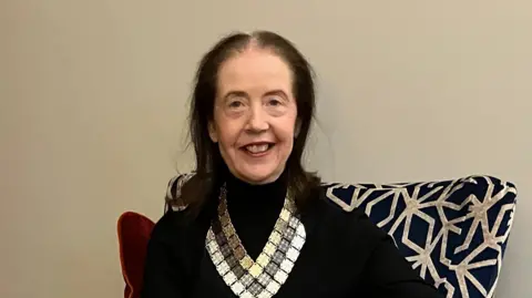 Karen Corrigan has long brown hair and is wearing a black long sleeved top with a jewelled lapel and a silver skirt. She is sitting on a black and white patterned armchair and smiling at the camera. 