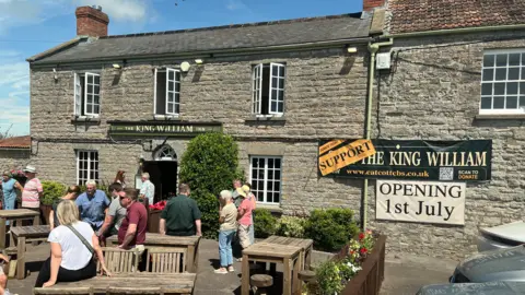 An old grey stone pub is pictured with a sign over the door which says 'The King William Inn'. There are also banners strung across the pub which say 'Support the King William - opening 1st July'. There are several people gathered outside the pub. Most are standing near some wooden tables and chairs.