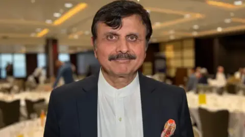 Dr Sarfaraz Jamali wears a white shirt and black blazer as he smiles toward the camera with an event including tables and guests taking place behind him