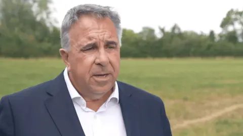 Steve Tuckwell in a green field surrounded by trees. He wears a navy suit jacket and a white shirt with an open collar.