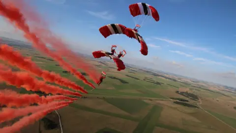 Red Devils Parachute Display Team Four men glide across a country landscape on red, white and black parachutes. Red smoke is billowing out behind them and they are choreographed directly above each other other as they descend