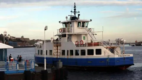 The Shields Ferry. The blue and white boat is pulling out from the dock. The boats name, Pride of the Tyne, is displayed at the rear end of the vessel.