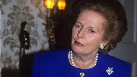 Margaret Thatcher has brown eyes and short brown hair swept backwards. She is wearing a cobalt blue collar-less blazer with a diamante brooch on the right collarbone, a pearl necklace and a pearl-stud earring. Her head is tilted towards the left and she is sitting in a room decorated with paisley-patterned wallpaper and there is a wall sconce, which is lit.