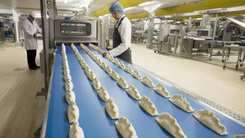 Wrights Four rows of pasties appear on a blue production line in a factory. A woman in protective uniform and hair netting stands over them coming out of a machine and a man stands to the left further away.