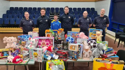 Supplied Luke stands in a line with five members of South Tyneside Fire and Rescue Service. In front of them are piles of children's toys arranged on tables. The toys include, dolls, dinosaurs, footballs and games.