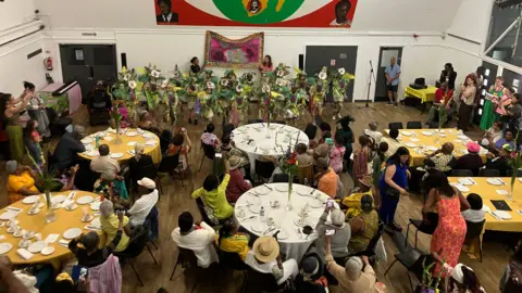 A parade of dancers wearing traditional carnival clothing are seen performing at the front of a large hall, where groups of people are watching and taking photos.