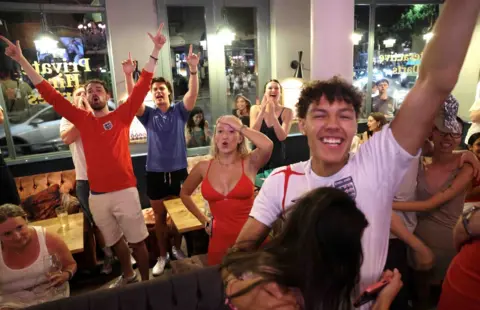 Getty Fans watch England's World Cup quarter-final against Norway at the Kings Arms pub in Fulham, south-west London. One woman clasps her hands together nervously while others stare at the screen. A St George's Cross headband and pints of beer sit on the table.
